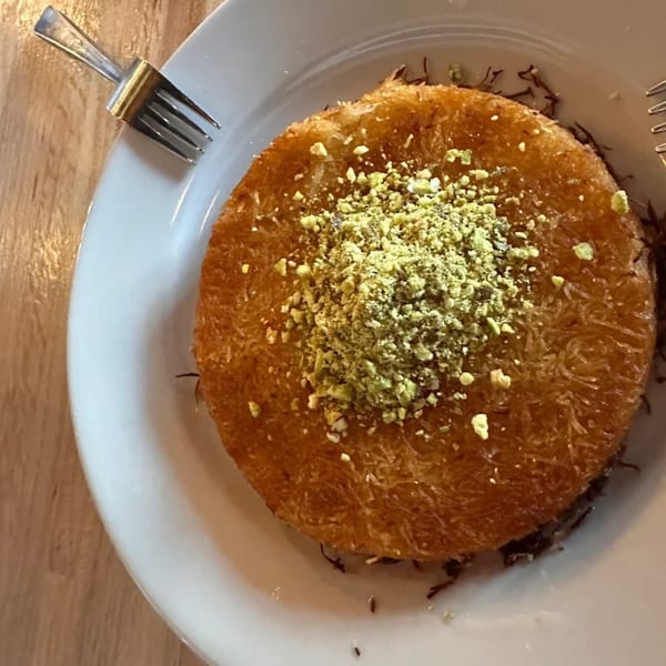Golden kunafa dessert topped with crushed pistachios on a white plate with a fork.