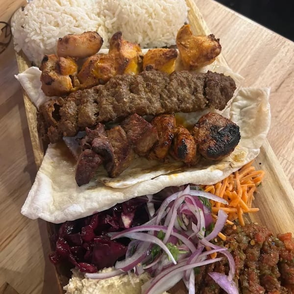 A wooden platter with rice, various grilled meats, flatbread, pickled vegetables, and hummus.
