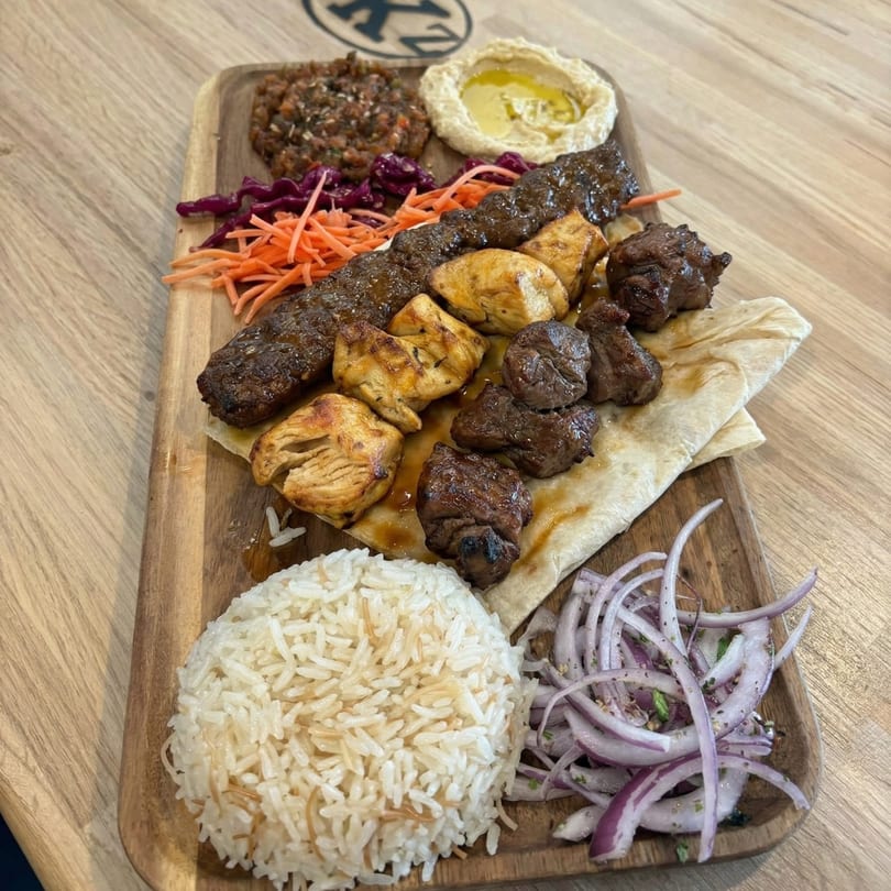 A wooden platter with a kebab skewer, chicken and beef chunks, rice, hummus, and salad.