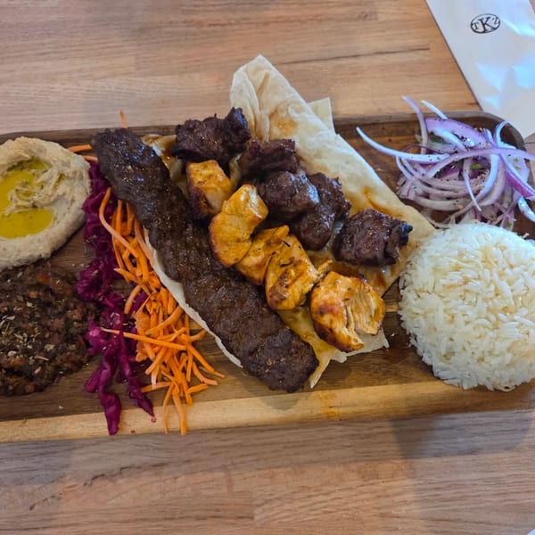 A wooden platter with various meats, rice, hummus, shredded carrots, red cabbage, and red onions.